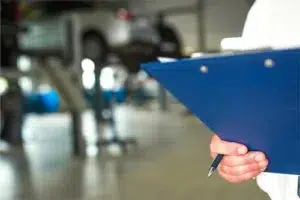 Are Local Auto Repair Shops Cheaper Than Dealerships? | ABS Unlimited in Fairfax, VA. Closeup image of an auto mechanic holding a clipboard for recording repairs and maintenance. The blurred background contains display of cars.
